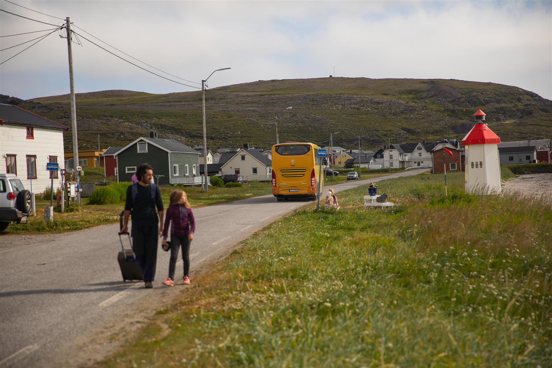 En småbarnfamilie fotografert langs en bygdevei, man kan se en gul buss ved en bussholdeplass. Det blåser, og bildet er tatt på sommeren.