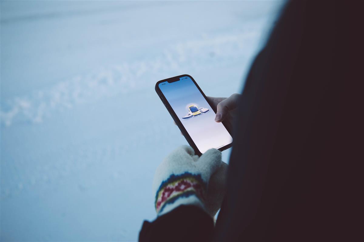 En person holder en mobiltelefon og på skjermen kan man se illustrasjoner av kollektivtransport. Personen har på seg en vott på den ene hånden, i forgrunnen kan man se personens rygg. Bildet er tatt på vinterstid. - Klikk for stort bilde