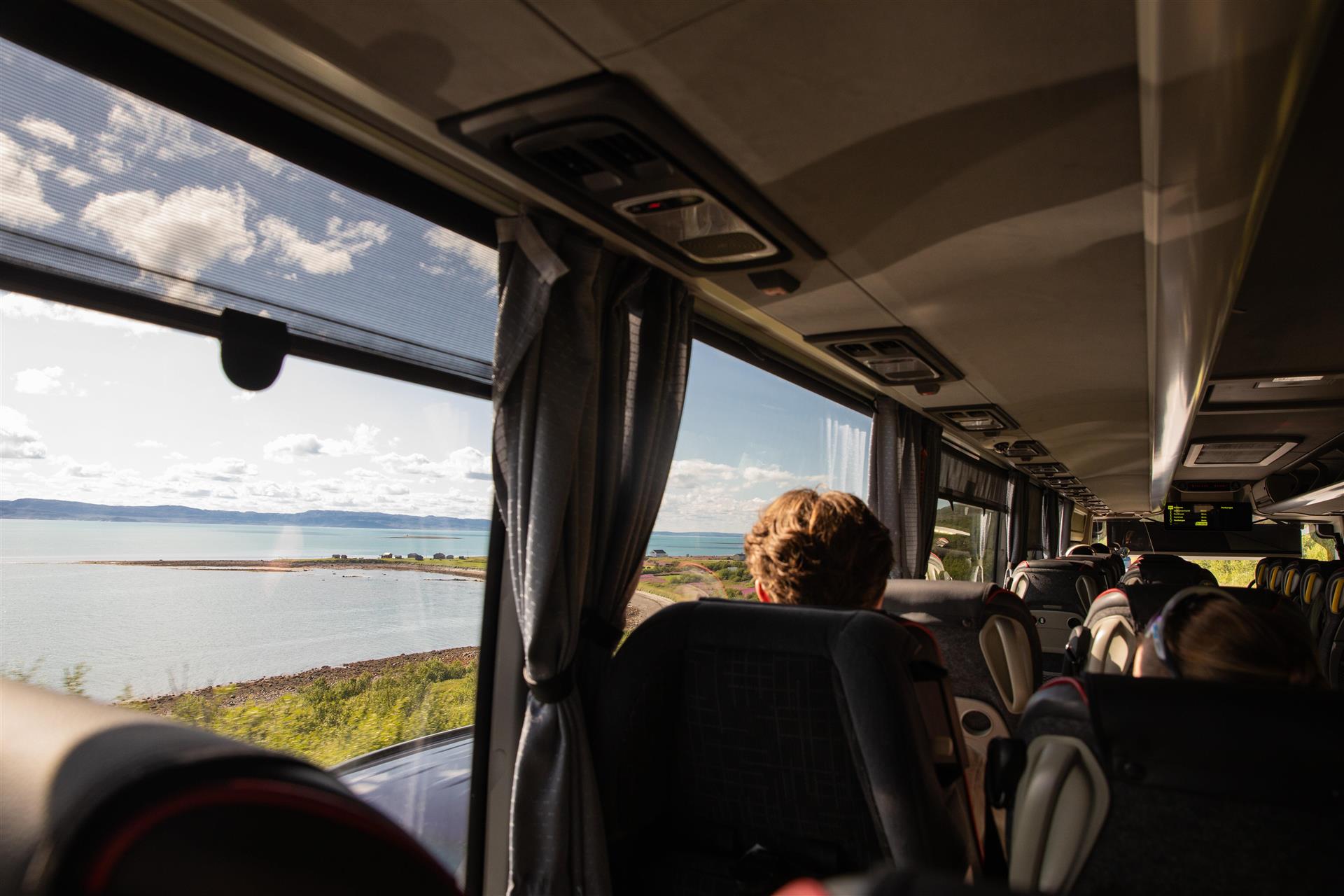 Bilde tatt inne fra en buss, man kan se flere passasjerer i sine seter, bildet er tatt en fin sommerdag, man kan se Varangerfjordlandskapet.