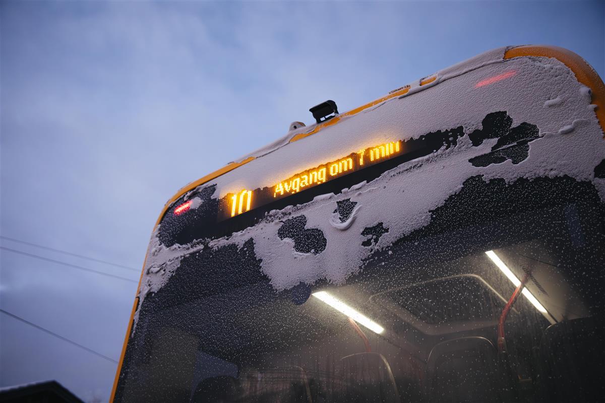 Baksiden av en gul buss. Det står "10 Avgang om 7 min" på infoskjermen. Bussen er full av snø. - Klikk for stort bilde
