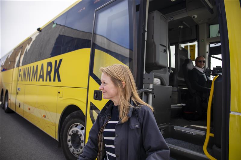 En ung, smilende kvinne går ut av en gul buss, på bussen står det "Finnmark" med stor skrift. I bakgrunnen ser man en smilende bussjåfør. Bildet er tatt på sommerhalvåret og sola skinner.