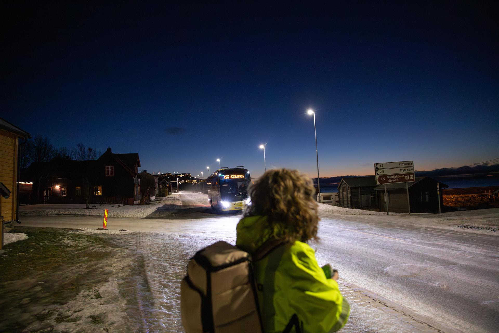 En kvinne i varselgul jakke holder en smarttelefon i hånden. Hun står ved en vei og ser mot en gul Snelandiabuss som kommer mot henne. Bildet er tatt på vinterstid på morgenkvisten.